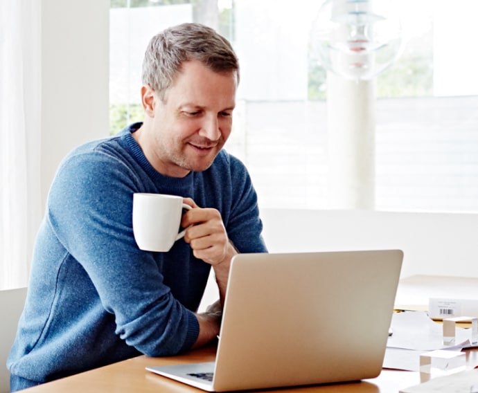 Άνδρας στο σπίτι χρησιμοποιεί φορητό υπολογιστή και πίνει καφέ.